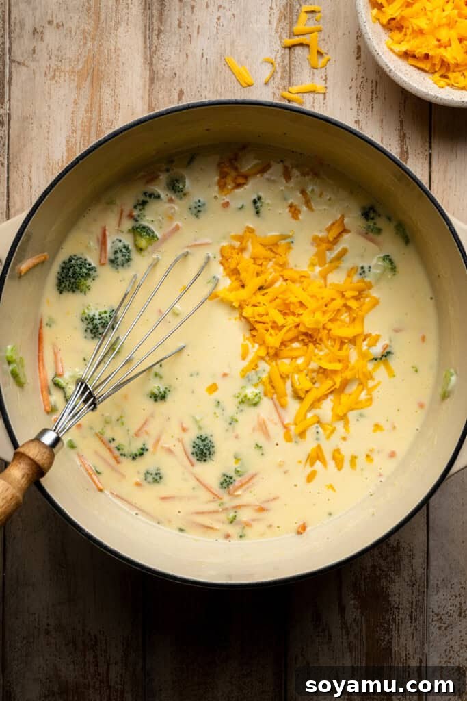 Freshly grated cheddar cheese being slowly whisked into the hot soup, melting to create a smooth, cheesy consistency.