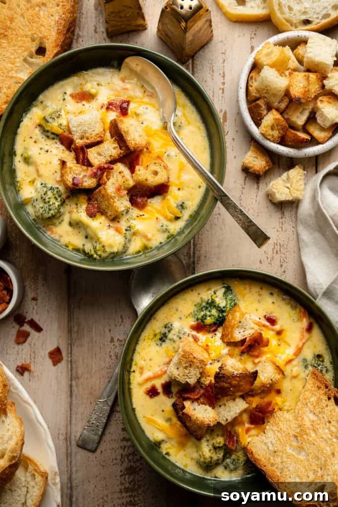 Two steaming bowls of creamy broccoli cheddar soup, garnished with crispy bacon and fresh herbs, sitting on a wooden table.