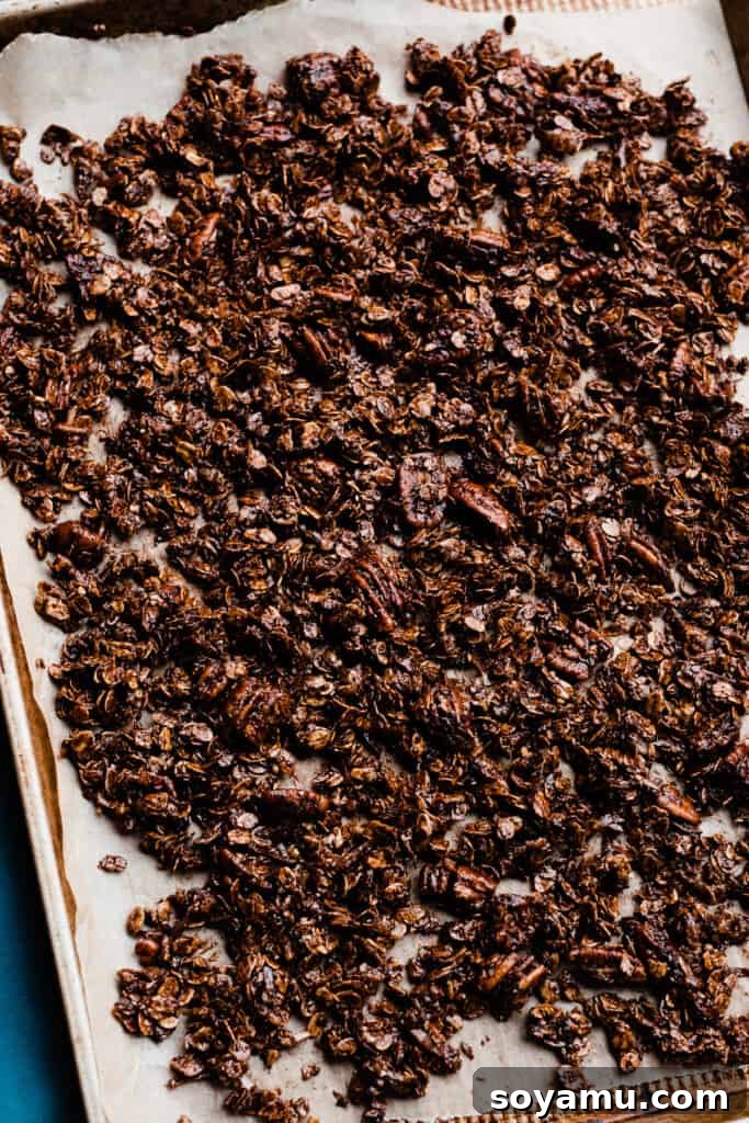 A sheet pan lined with parchment paper, evenly spread with the unbaked chocolate granola mixture, ready for the oven.