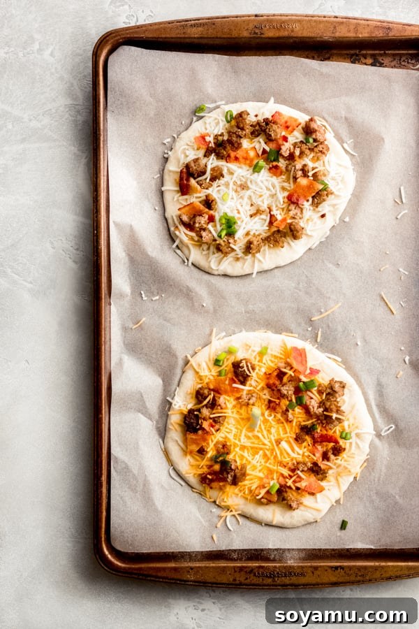 Individual breakfast pizzas arranged on a baking sheet, ready for the oven. Several raw breakfast pizzas assembled on a baking tray, topped with cheese and meat, prior to baking.