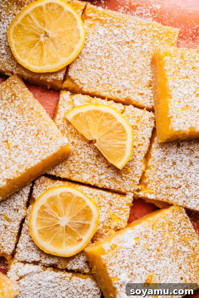 Effortless Zesty Lemon Bars 3 A close-up shot of perfectly sliced lemon bars arranged on a delicate pink surface. Each bar is generously dusted with powdered sugar, and a few are garnished with fresh lemon slices, highlighting their vibrant citrus essence.