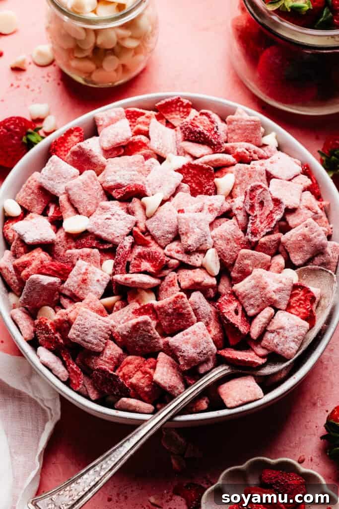 4-Ingredient Strawberries and Cream Puppy Chow Recipe 3 A bowl of strawberry puppy chow on a pink surface, surrounded by a cup of white chocolate chips and some fresh strawberries.