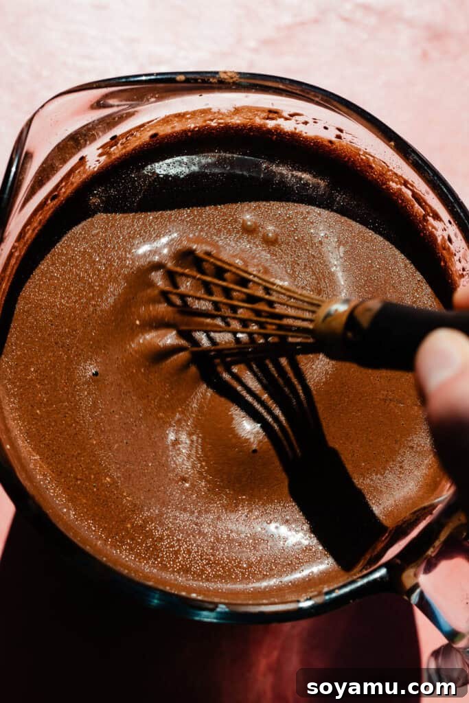 A whisk stirring the bloomed cocoa mixture in a measuring cup.