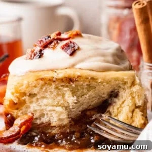 A close-up of a maple bacon cinnamon roll on a plate, with the maple caramel pan sauce visible underneath the roll.